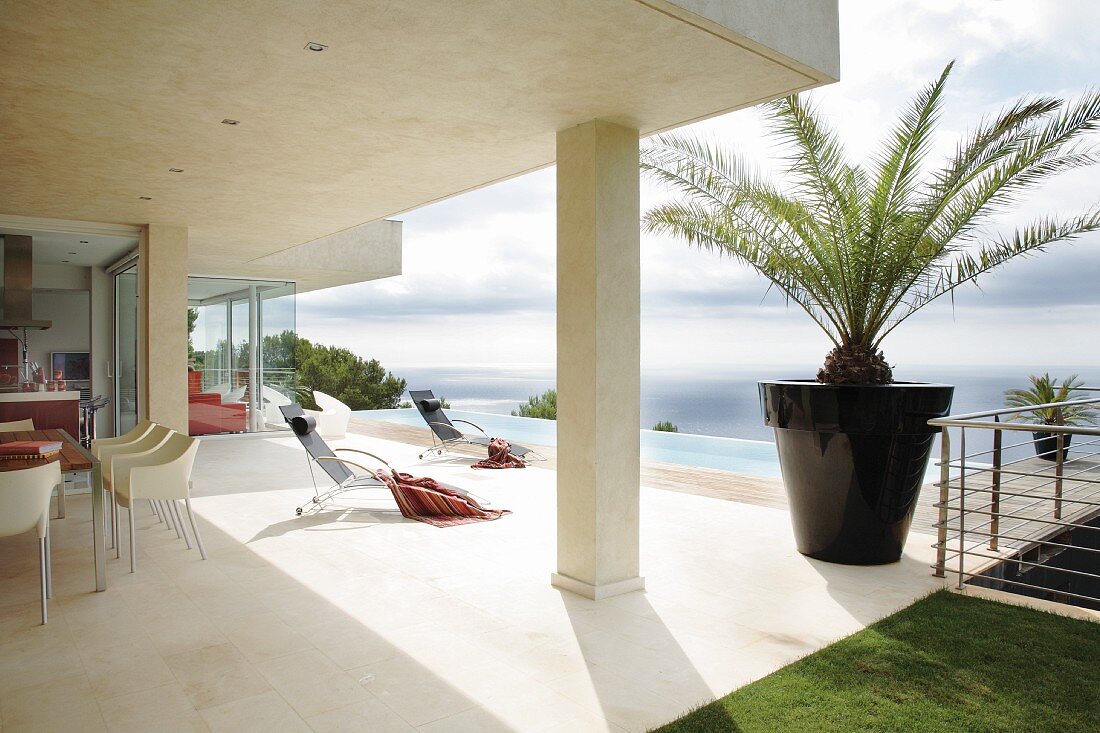 Designer dining area below projecting concrete roof and sun loungers on pool terrace of modern holiday home by the sea