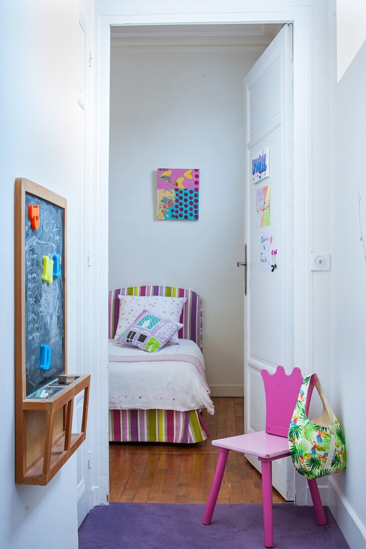Pinkfarbener Kinderstuhl gegenüber Wandtafel in kleinem Vorraum mit offener Tür und Blick auf Bett mit gestreifter Husse