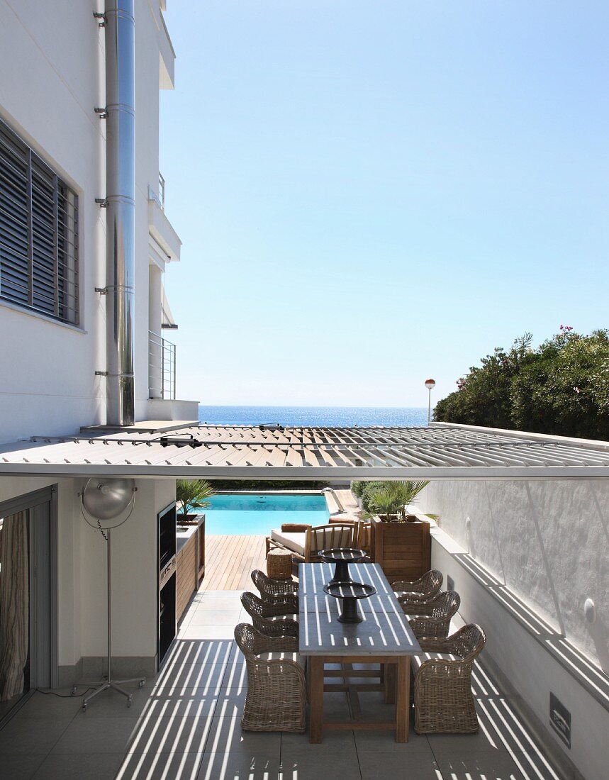 Mit Sonnenschutzlamellen überdachte Terrasse zwischen mehrstöckigem Wohnhaus und Gartenmauer, im Hintergrund das Meer