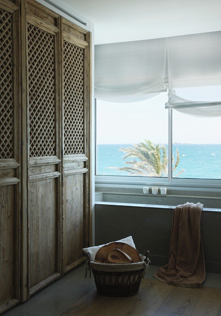 Einbauschrank mit rustikalen Holztüren teilweise aus Holzgitter, neben Badewanne am Fenster mit Meerblick