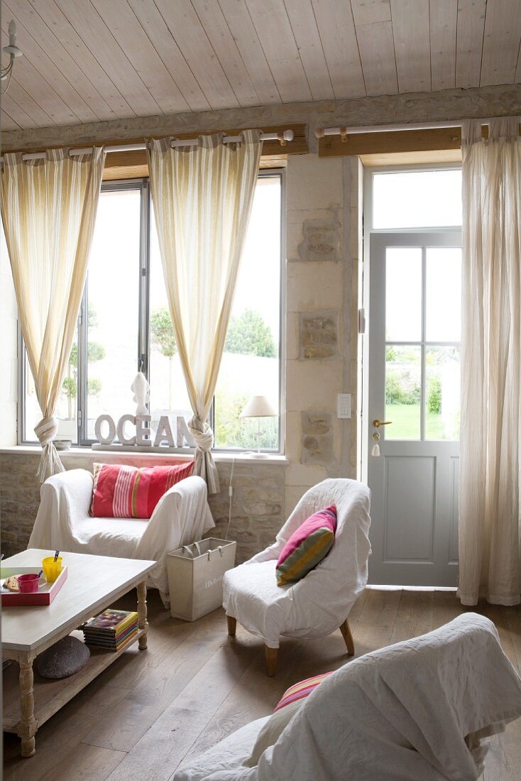 Various armchairs with white loose covers around coffee tables in front of large window with knotted curtains in simple living room