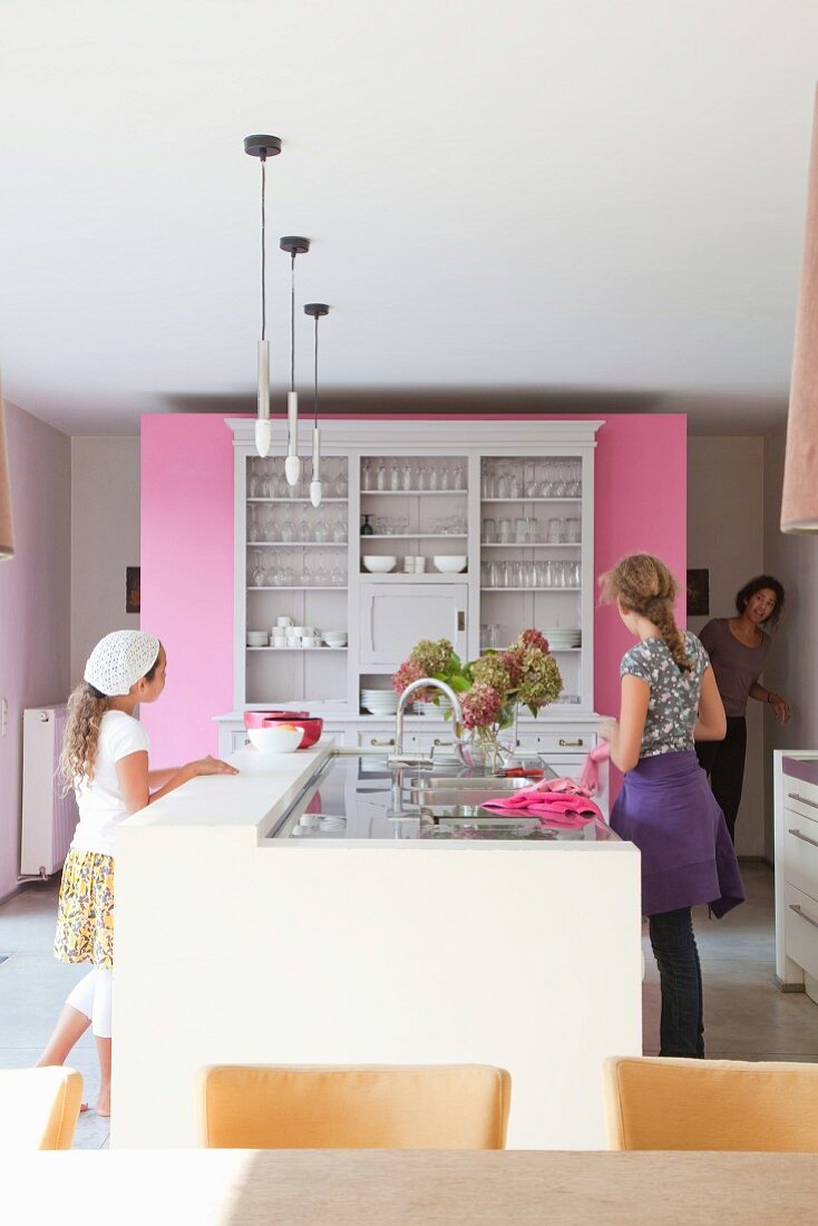 Moderne Küchentheke vor Vintage-Geschirrschrank an pinkfarbener Raumteilerwand, zwei Mädchen und Frau im Hintergrund