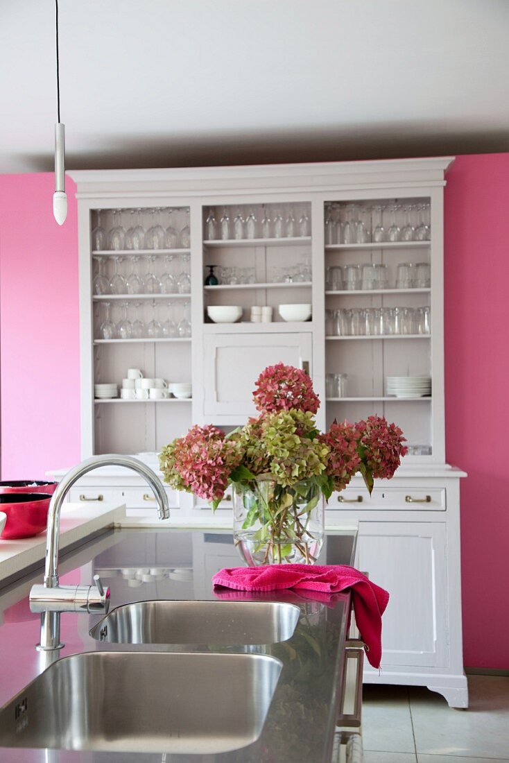 Collection of glasses in pale, vintage dresser against pink partition wall in open-plan kitchen