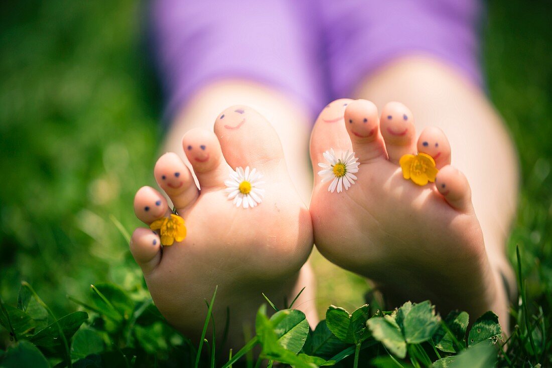 Mit Wiesenblumen (Gänseblümchen, Butterblume) und Smileys geschmückte Füsse eines im Gras liegenden Mädchens