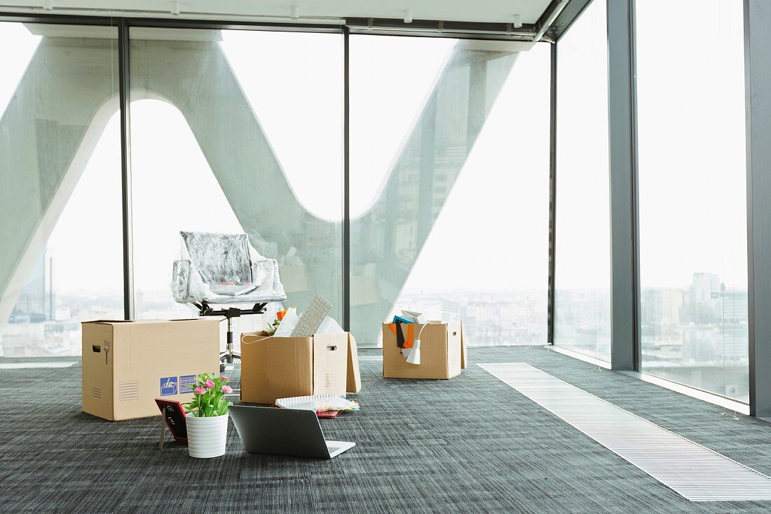Umzugskisten, offener Laptop auf dem Boden und Bürostuhl in leerem Hochhausbüro mit Glasfassade