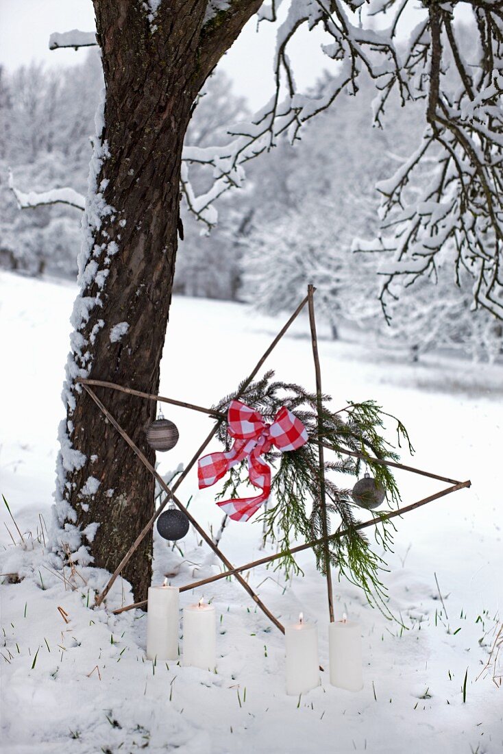 Dekostern aus Holzstäben am Baum lehnend, mit Weihnachtskugeln und Schleife geschmückt, davor brennende Kerzen im Schnee