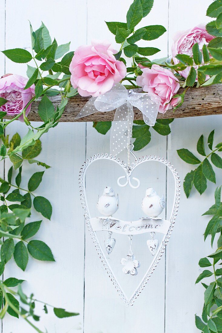 Heart-shaped, ornate metal welcome sign hanging from branch decorated with garland of roses