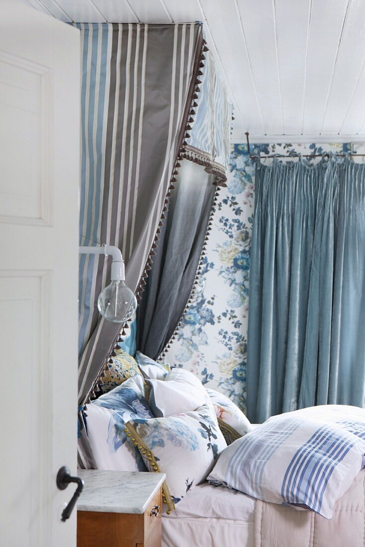 Half-tester bed with many scatter cushions and blue and grey striped canopy against floral wallpaper in romantic bedroom in shades of blue