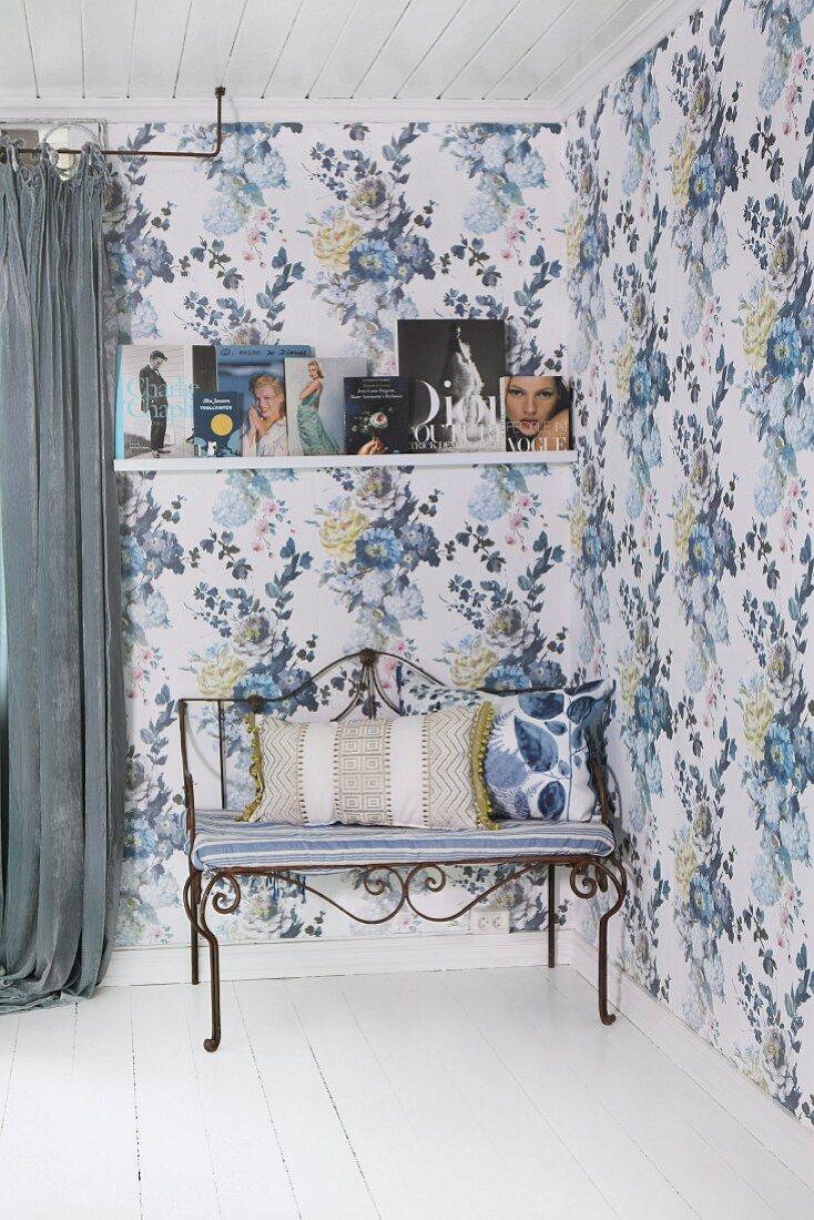 Delicate metal bench with seat cushion and scatter cushion in corner of romantic room with blue floral wallpaper