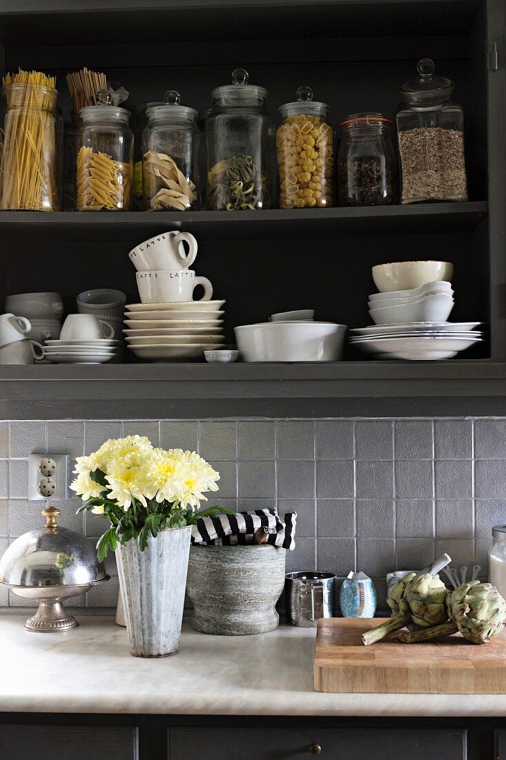 Blumenstrauss auf Küchenzeile, darüber offener Hängeschrank mit weißem Geschirr und Vorratsgläsern