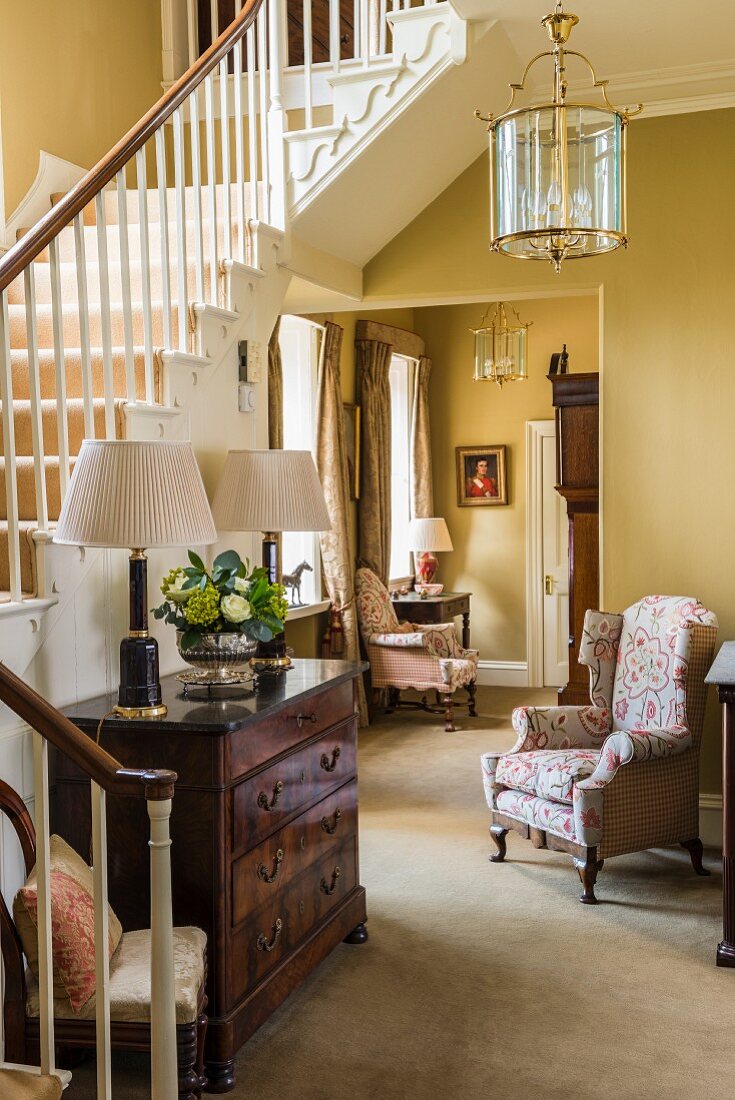 Flurbereich im klassischen Stil mit antiker Kommode unter der Treppe und Sessel mit floralem Bezug
