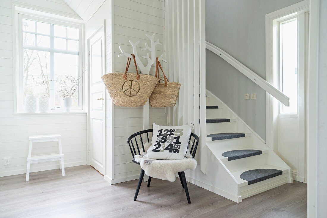 White, wood-clad foyer with step ladder below window, chair and tree-shaped coat rack next to foot of staircase with grey runner