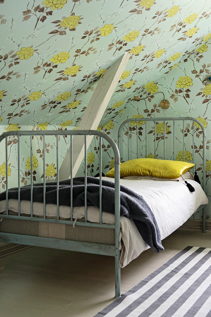Grey-painted vintage bed under sloping ceiling with vintage-style floral wallpaper