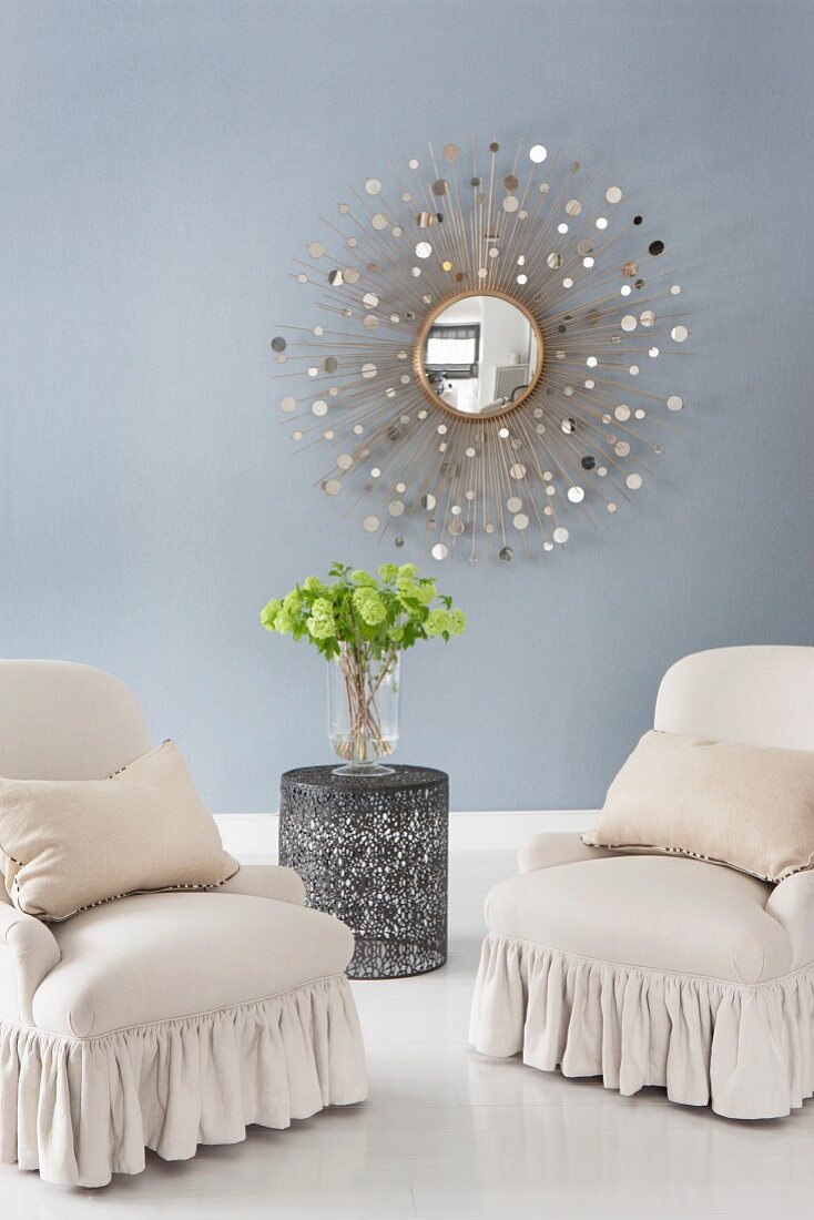Elegante Polstersessel mit Volant auf weiß lackiertem Dielenboden und an blauer Wand Wandspiegel in Strahlenform