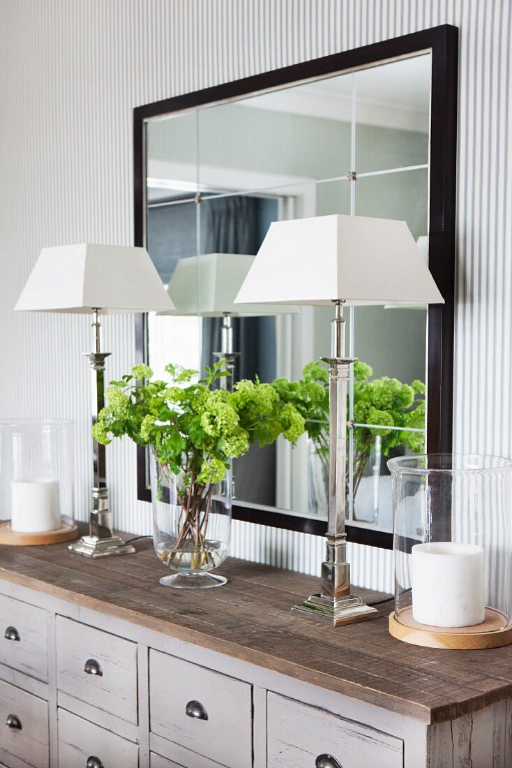 Elegante Silber-Tischleuchten mit weißem Lampenschirm auf grauer Apothekerschrank und vor schwarz gerahmtem Wandspiegel in edlem Ambiente