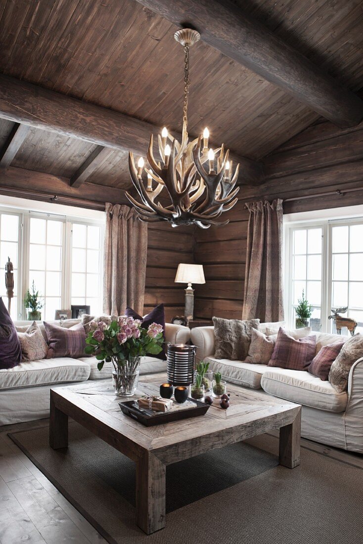 Couchtisch aus Massivholz und helle Sofagarnitur unter Geweih-Leuchte in Wohnzimmerecke eines Holzhauses