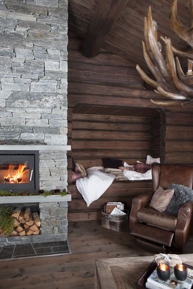 Cosy living area with brown leather armchair in front of alcove in log wall and fire in fireplace