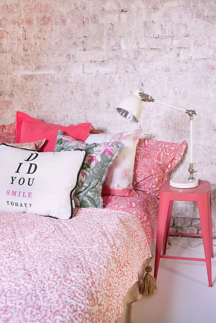 Bedside lamp on pink-painted retro-style metal stool next to bed with red and white patterned bedspread and scatter cushions against brick wall