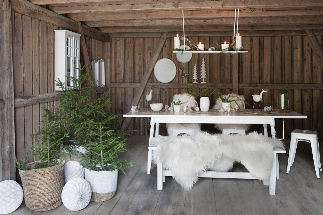 White table and bench with white sheepskins next to small potted Christmas trees in wooden house