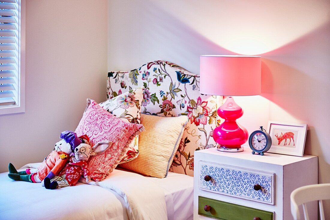 Stofftiere und Kissen auf Bett mit Kopfteil und Blumenmuster auf Stoffbezug, daneben Tischleuchte in Rosa auf Nachttisch