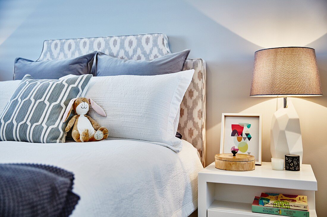 Scatter cushions and rabbit soft toy on double bed with upholstered headboard; table lamp on white bedside table