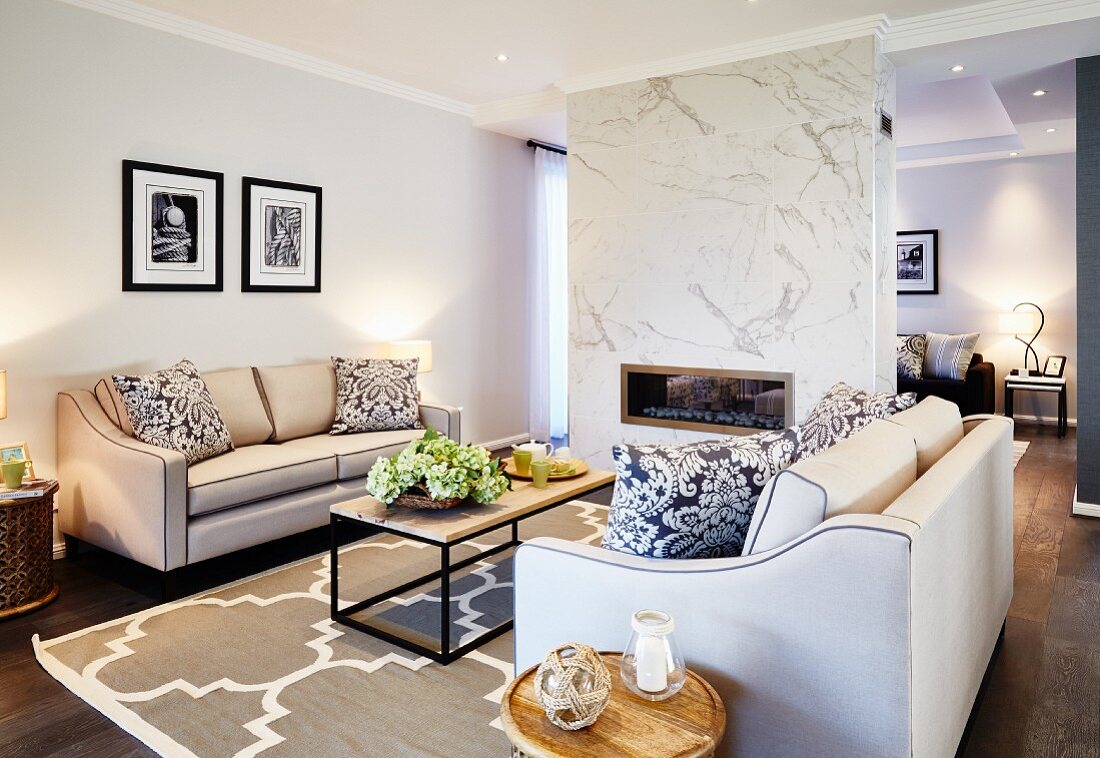 Elegante Sofagarnitur mit hellem Bezug und moderner Couchtisch auf Teppich, mit grossformatigem Ornament Muster in offenem Wohnraum, Kamin in Raumteiler