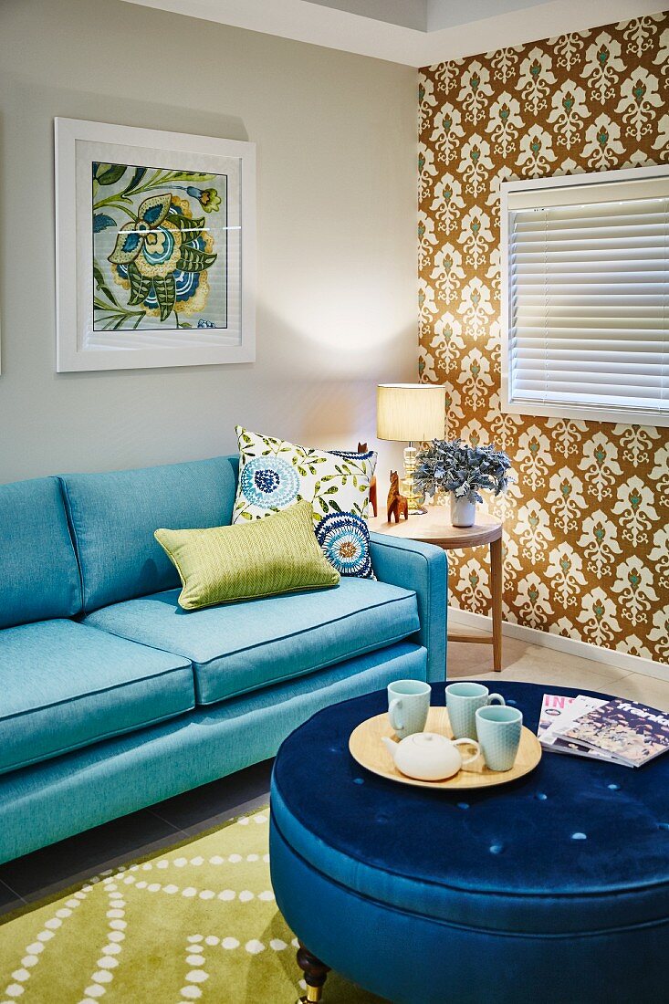 Tablett mit Tassen auf rundem Polstertisch mit blauem Satinbezug und hellblaues Sofa in Wohnzimmer, seitlich tapezierte Wand mit Retro Ornament Muster