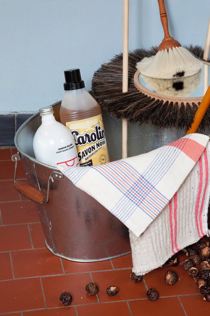 Bottles of cleaning agent, cleaning cloths and special brushes in zinc bucket; soap nuts on tiled floor