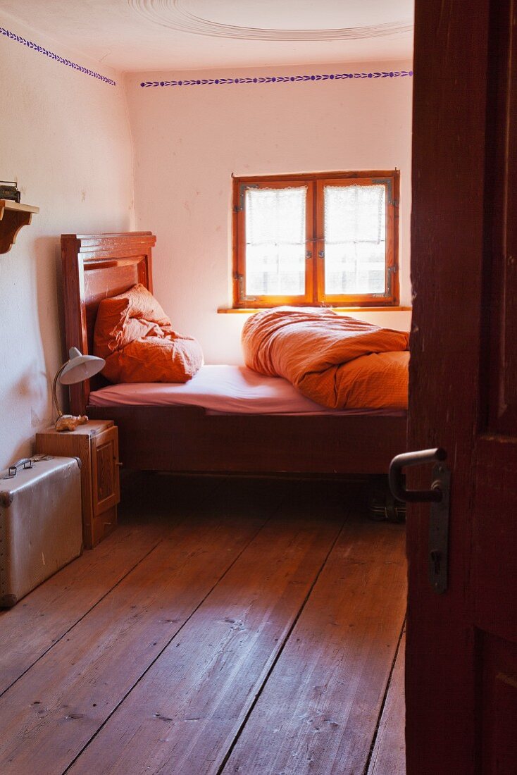 Blick durch offene Tür in einfaches Bauern-Schlafzimmer mit traditionellem Holzbett vor Fenster
