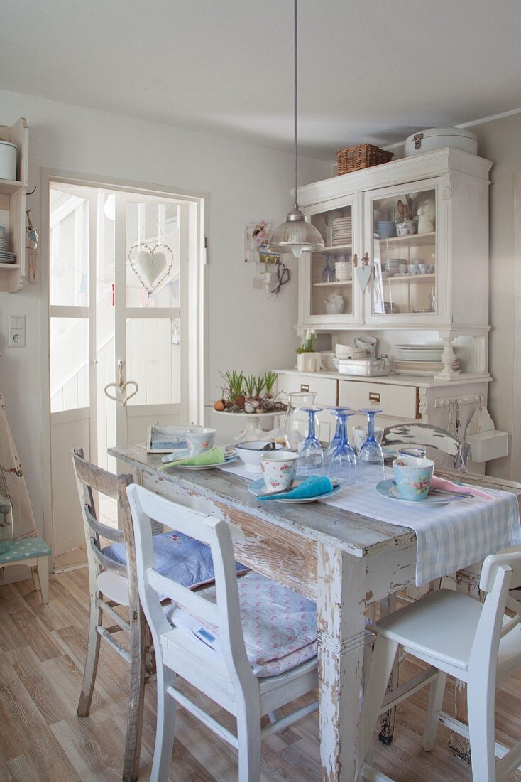 Pastellfarben gedeckter Shabby Esstisch mit alten Küchenstühlen, antiker, weisser Geschirrschrank im Hintergrund