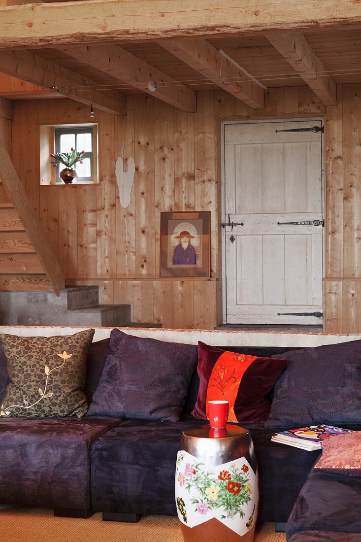 Tonnenförmiger Beistelltisch vor gemütlichem Polstersofa mit dunkelviolettem Bezug und Kissen, in Wohnraum mit Holzverkleidung