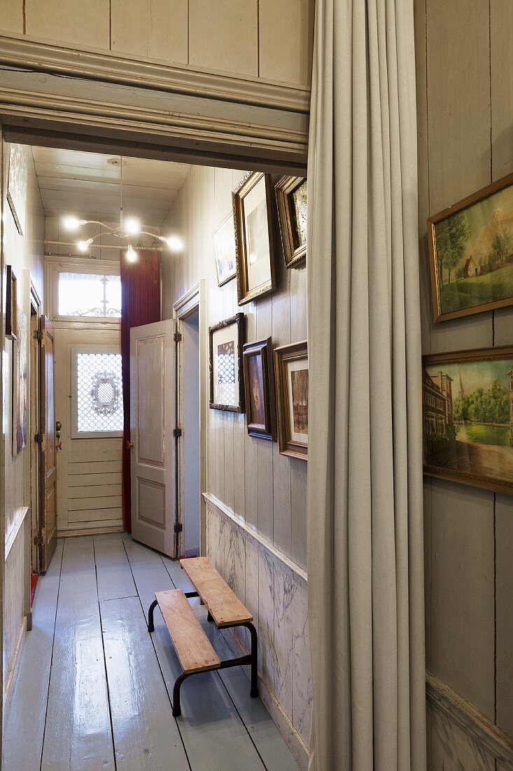 Narrow hallway with white-painted wooden floor, gallery of pictures and pale curtain in foreground