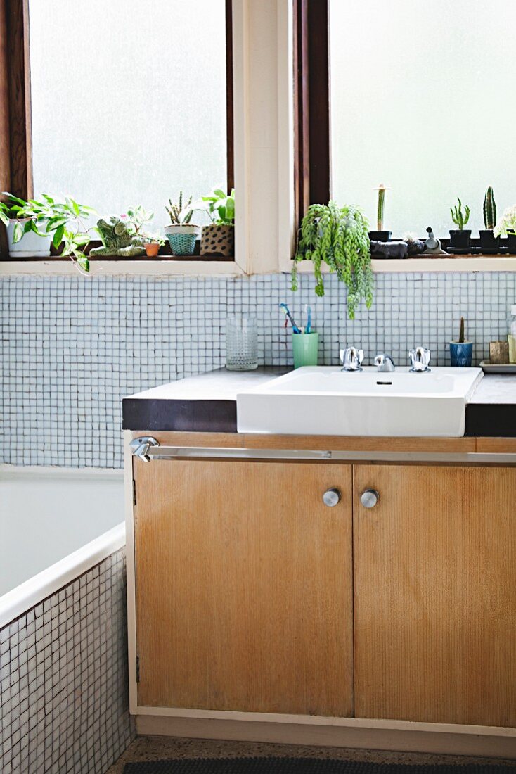 Retro-Waschtisch aus hellem Holz mit eingebautem Waschbecken vor Fenster, helle Mosaikfliesen an Wand und Badewanne
