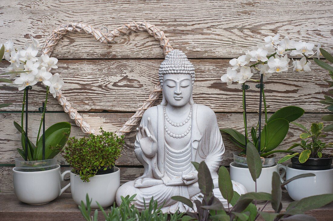 Potted herbs and white orchids next to Buddha statue in front of white love-heart hung on wooden wall