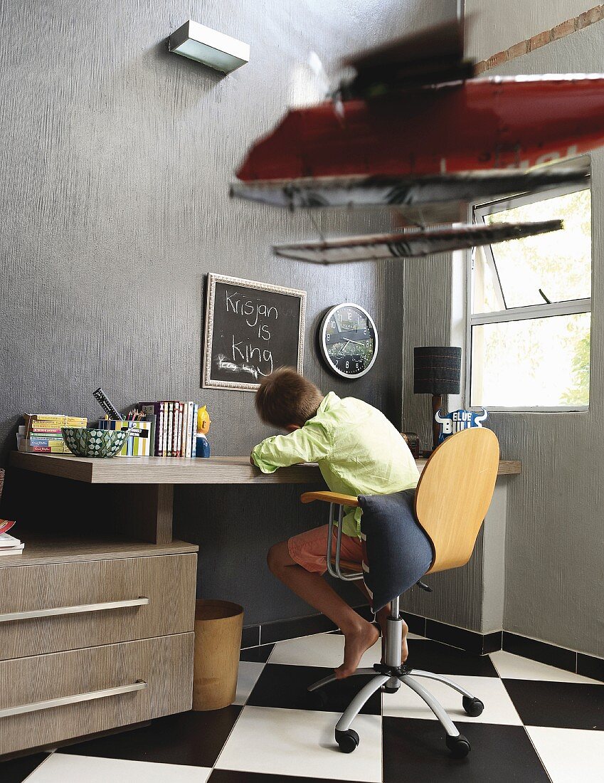 Boy sitting on swivel chair at desk in bedroom with chequered tiled floor
