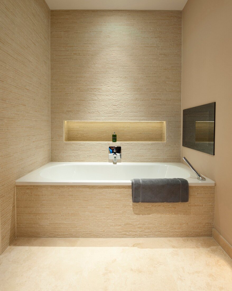 Fitted bathtub in designer bathroom with beige strip tiles; shelf niche with indirect lighting in back wall