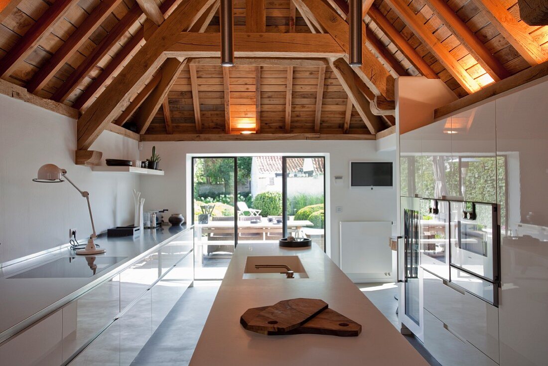 weiße Hochglanz-Designerküche unter rustikaler Holzbalkenkonstruktion und Blick auf sommerliche Terrasse