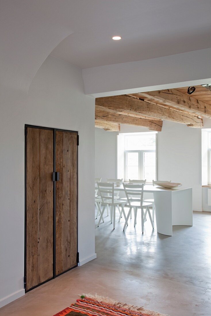 Minimalist, white dining area below rustic wooden ceiling beams in renovated interior