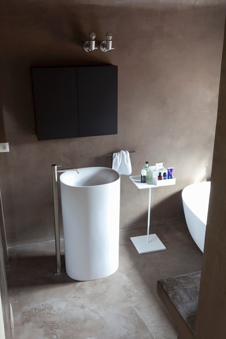 White pedestal sink with standpipe tap, black wall-mounted cabinet and concrete floor in minimalist designer bathroom