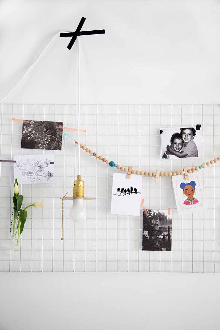 Weißes Metallgitter als Pinnwand mit Fotografien und Perlenschnur dekoriert, davor reduzierte Pendelleuchte mit Masking Tape festgeklebt
