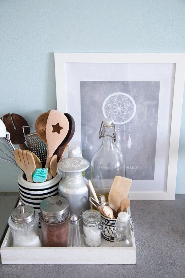Still-life arrangement of various retro kitchen utensils on white wooden tray in front of picture of dream-catcher