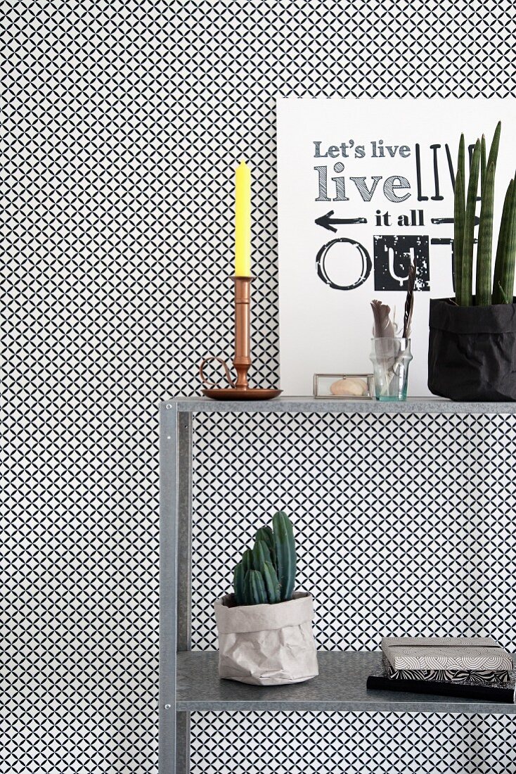 Still-life arrangement of black and white graphic artwork and yellow candle on metal shelves against black and white wallpaper