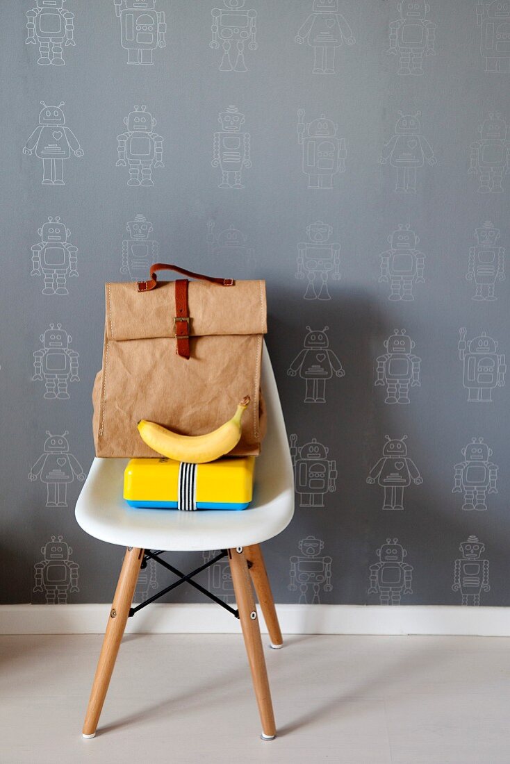 Tasche und gelbe Pausenbox mit Banane auf weißem Eames Chair vor grauer Tapete mit kindlichen Roboterfiguren