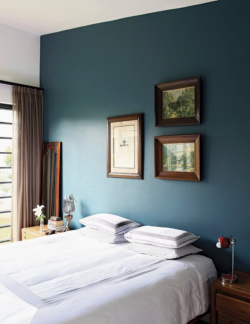 Bed with white bed line below framed pictures on petrol-blue bedroom wall