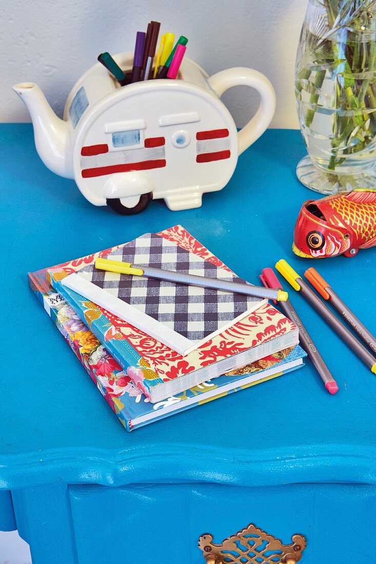 Colourful exercise books and felt-tip pens in front of retro teapot used as pen holder on blue table