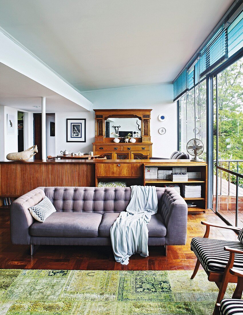 Hellgraue Couch vor Sideboard aus Holz, im Hintergrund Kommode mit Spiegelaufsatz, seitlich Fensterfront und offene Tür