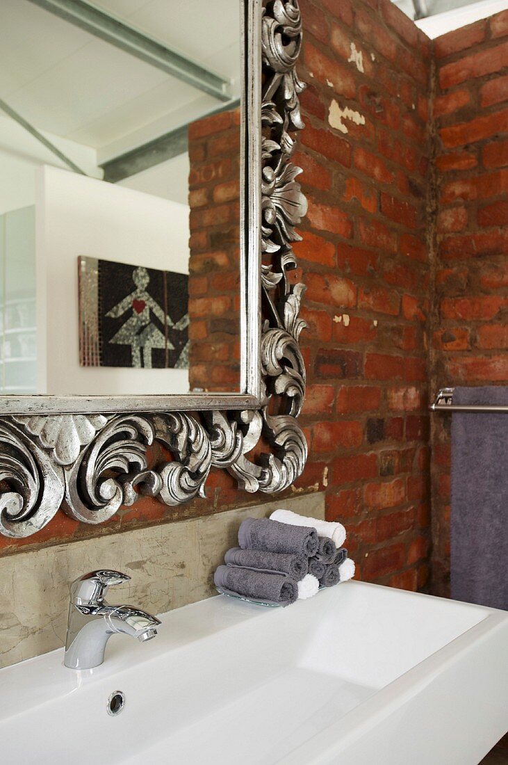 Mirror with ornate, floral silver frame and trough-style sink on brick wall