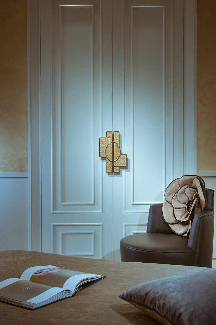 Blick über Bett auf weiße Zimmertür mit Profilleisten und dekadenter Türöffner, seitlich brauner Designer-Ledersessel