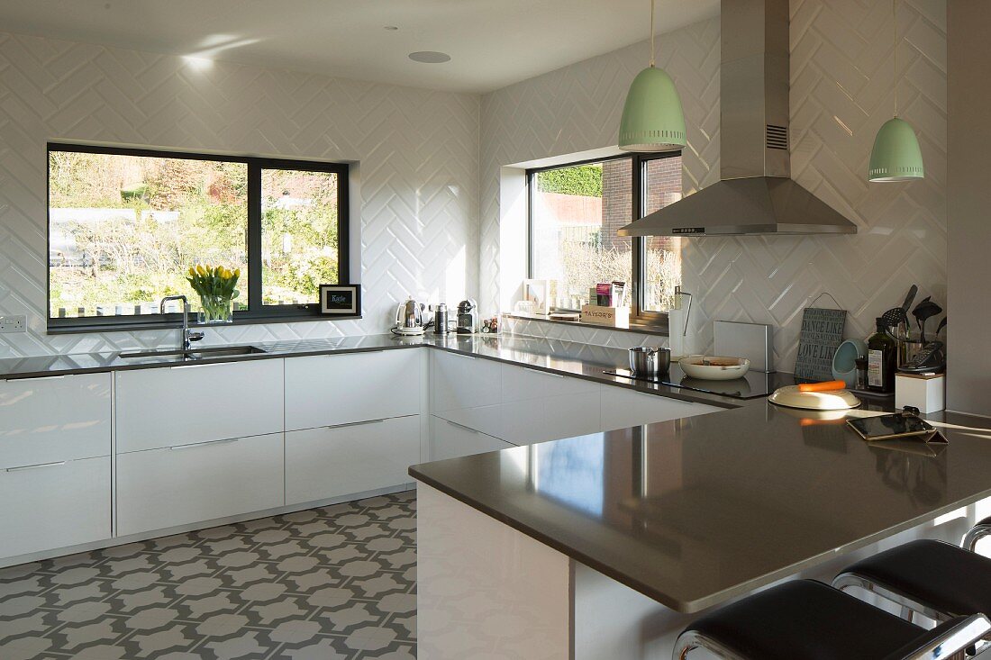 U-shaped, white, designer fitted kitchen with glossy worksurface below windows; breakfast bar and bar stools in foreground