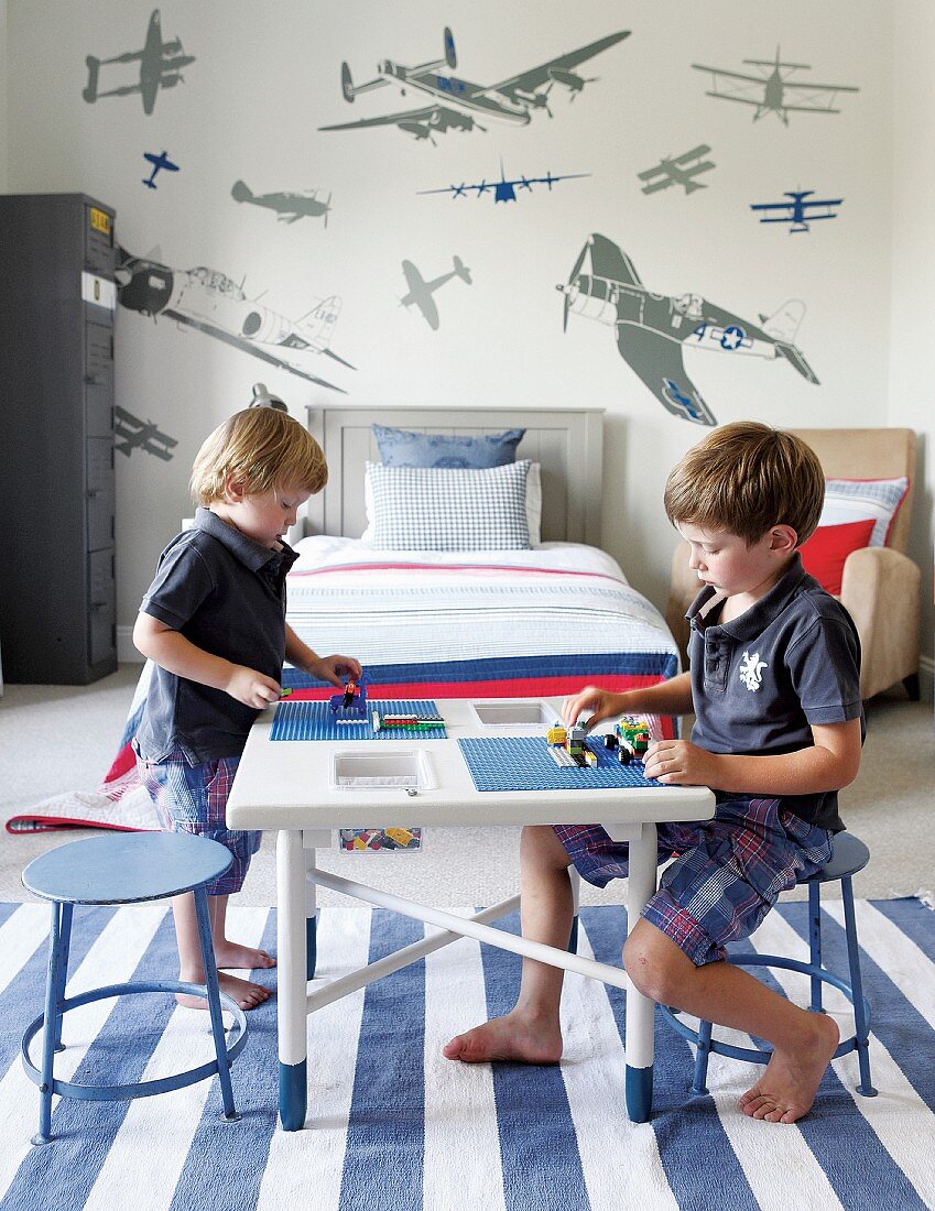 Two children playing on table in child's bedroom on blue and white striped rug; aeroplane wall sticker in background